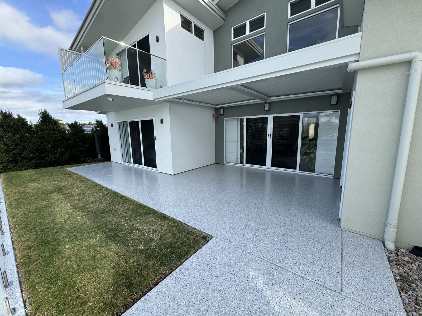 Large outdoor alfresco area finished in light grey flake epoxy, running from the lawn up to the sliding doors of a two-storey rendered home