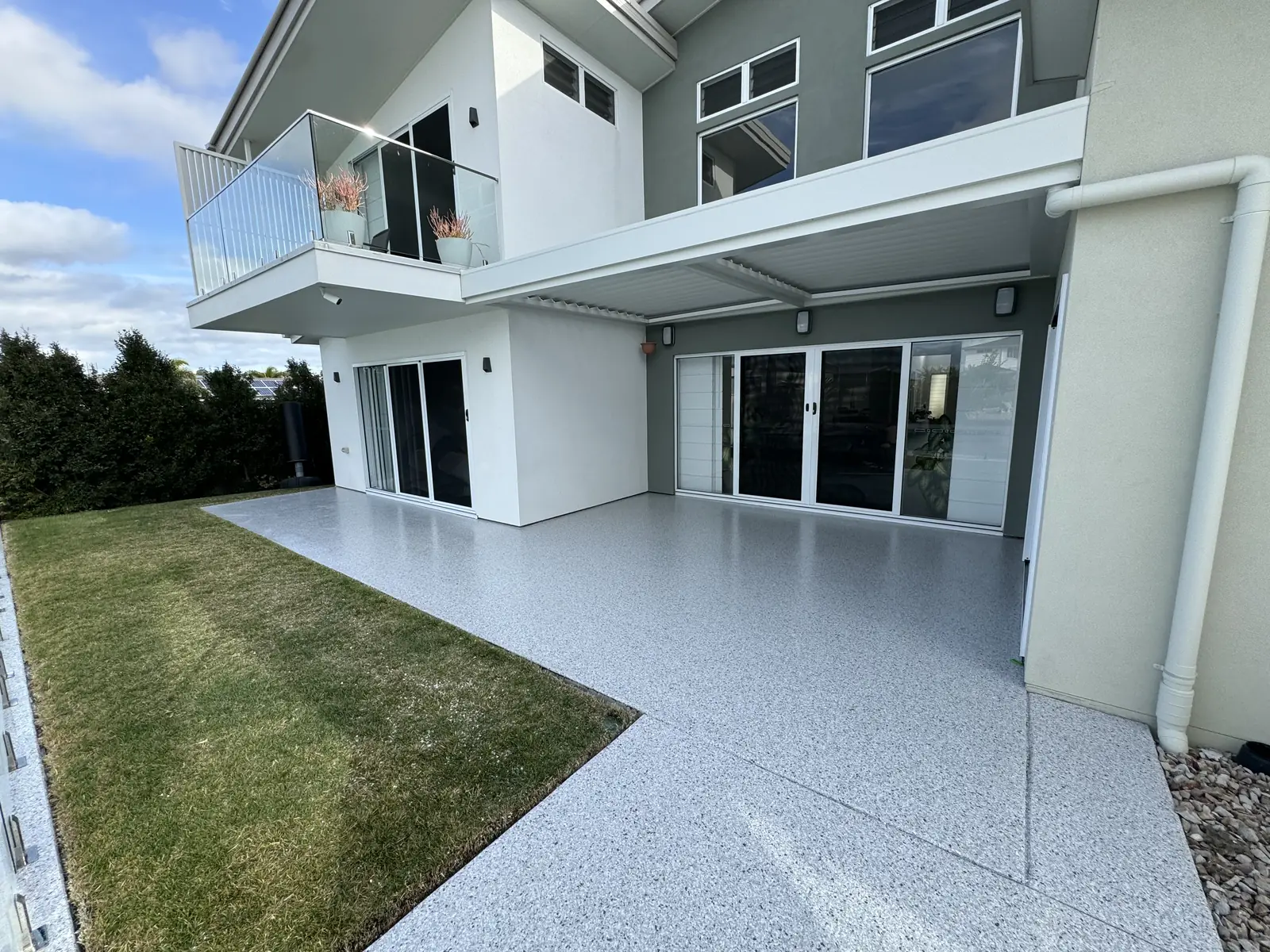 Large outdoor alfresco area finished in light grey flake epoxy, running from the lawn up to the sliding doors of a two-storey rendered home