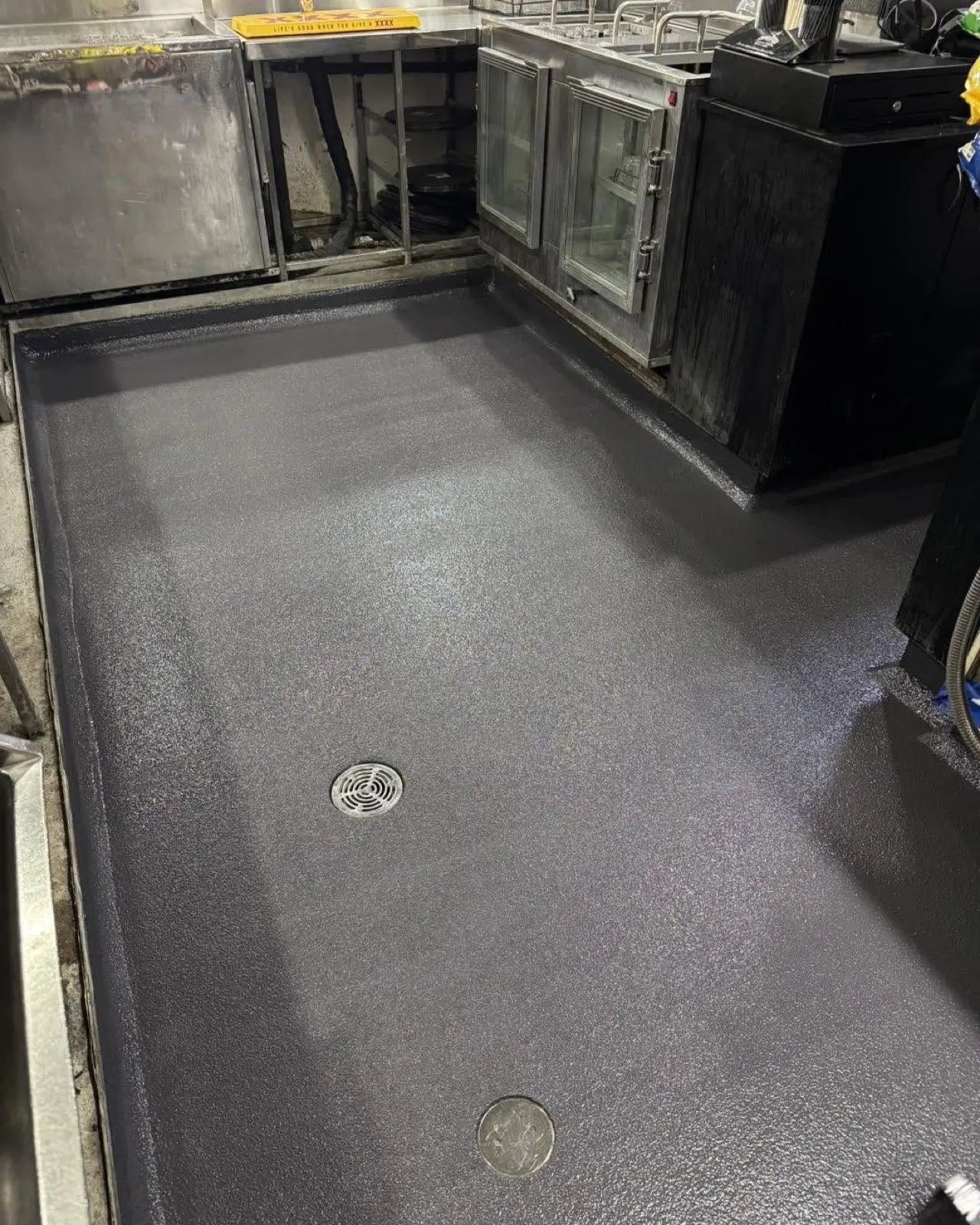 Dark grey non-slip epoxy floor in a commercial kitchen, coved up to the walls with a stainless floor waste between the benches