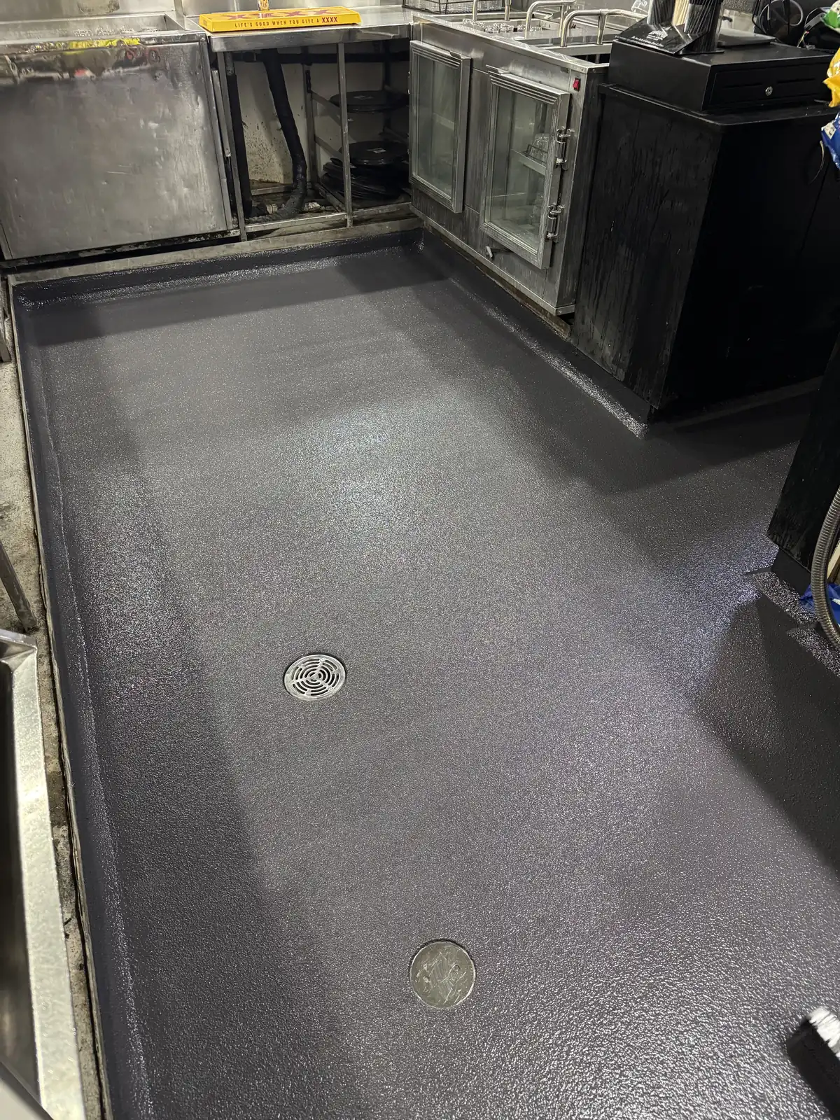 Dark non-slip hi-impact epoxy floor behind the counter of a commercial kitchen and bar, coved at the edges with two floor wastes